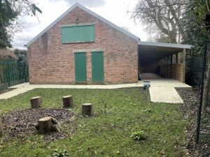 A photo of a Scout building with a large outdoor covered area to the right. In the foreground is a pathway with a ramp, leading to a clear and tidy grassy area. On the left hand side is tree stump seating around a fire pit.