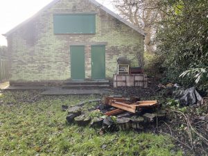 Photo of an old Scout building and an outside area, with overgrown bushes and brambles. In the foreground is an unkept grassy area with a fire pit circle.