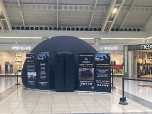 A photo of the UK Astronomy inflatable planetarium in situ at an event in a shopping centre. The planetarium is a black dome shape with a rectangle entrance.