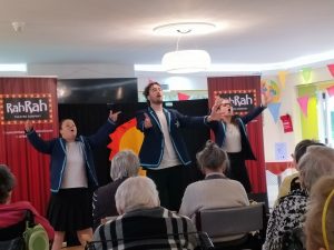 Three people dressed in blue coats singing to a small audience of elderly people.