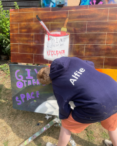 A young man with a blue hoodie with the name Alfie on the back is painting pictures, including one that says "Paint a life of non violence".