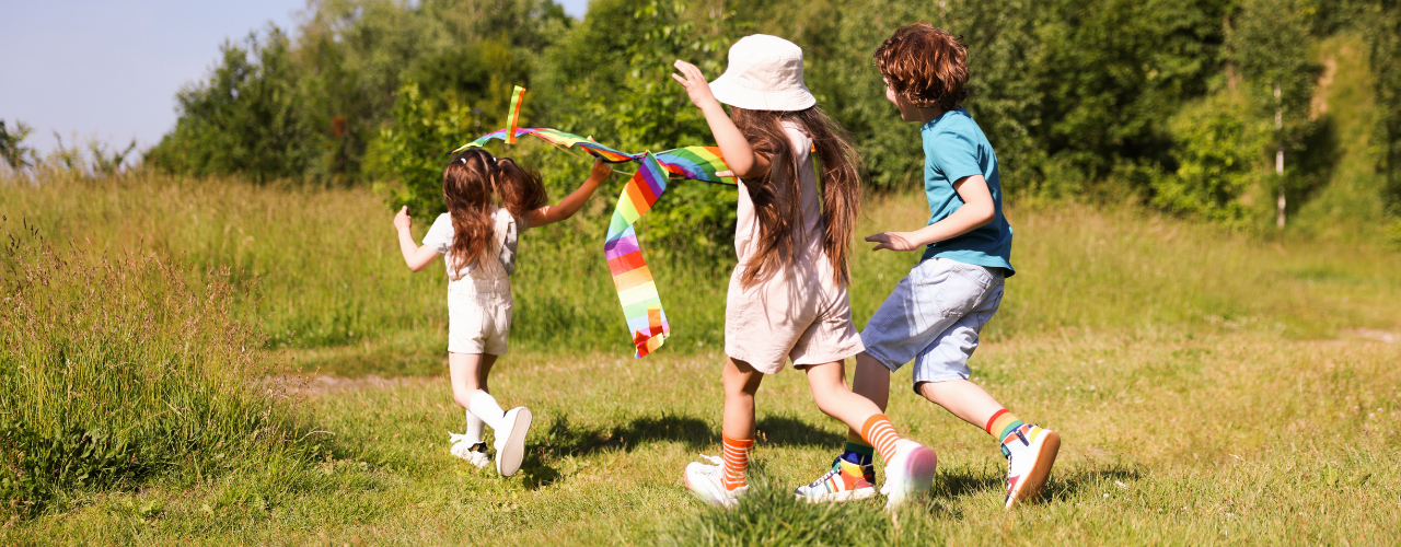 three children running across a grassy area carrying a colourful kite